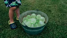 Vattenballonger ligger i en plåtbalja och ett barn står bredvid
